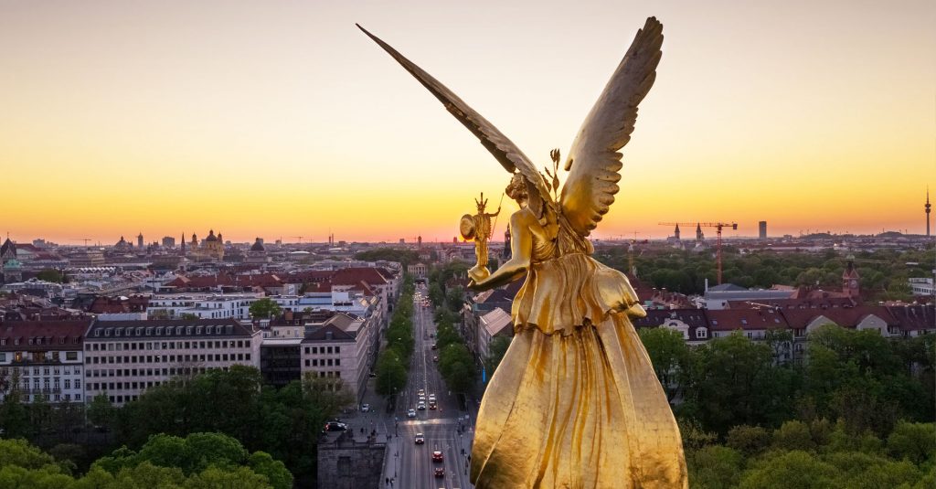 Eine Statu von einem Goldenen Engel. Im Hintergrund eine Großstadt mit Mehrfamilienhäusern und Kirchen.